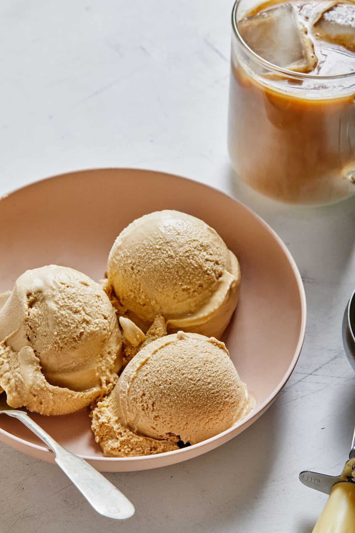 Three scoops of ice cream on top of a plate with a spoon and a glass of coffee beside it