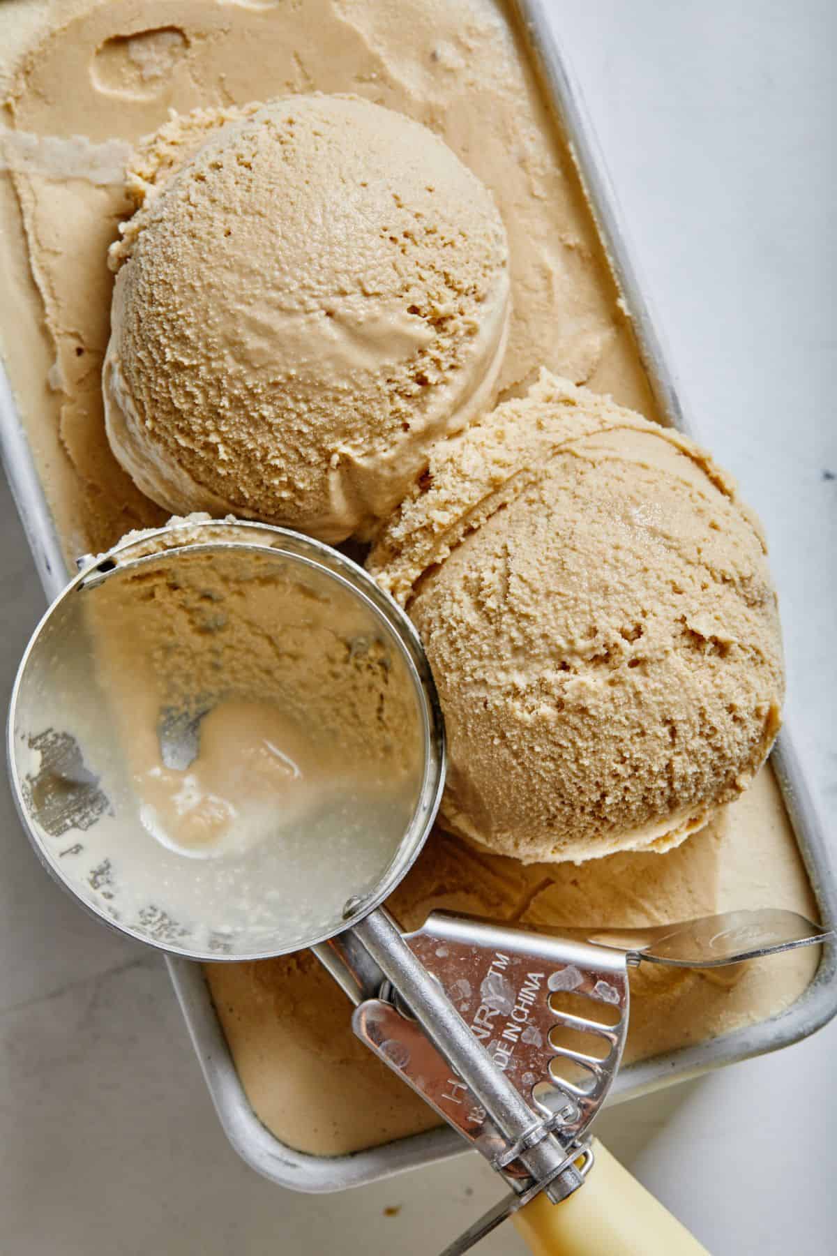Two scoops of coffee ice cream resting on top of an ice cream tub with an ice cream scoop beside it
