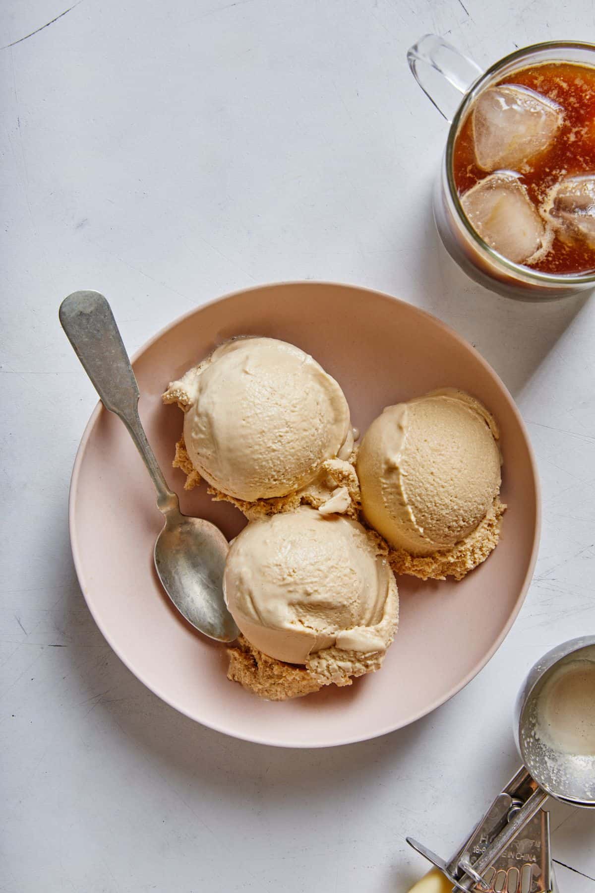 Three scoops of ice cream on top of a plate with a spoon and a glass of coffee and an ice cream scoop beside it