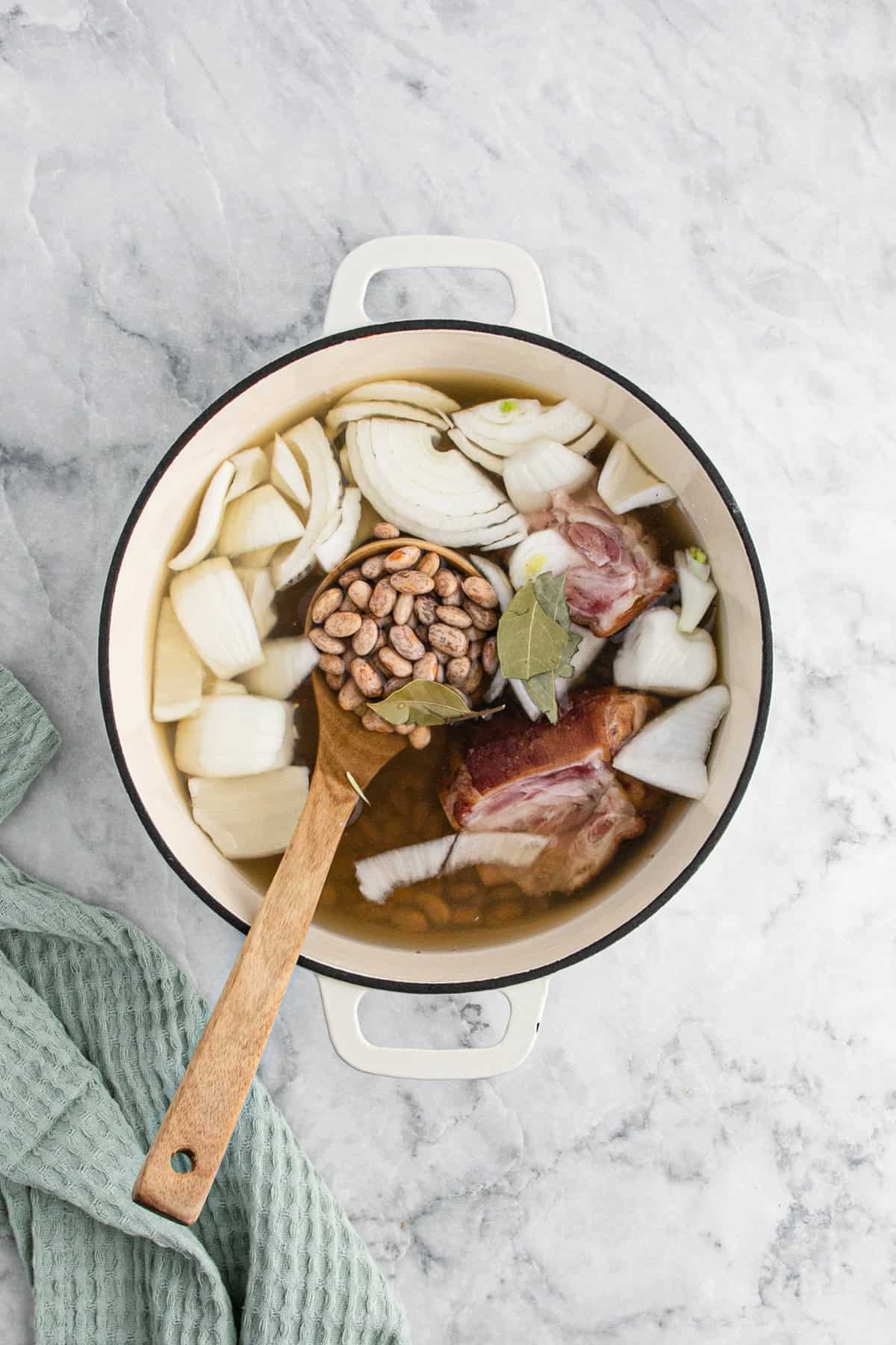 Pinto beans, chicken stock, seasonings, onion and spices in a large pot being stirred together on white countertop