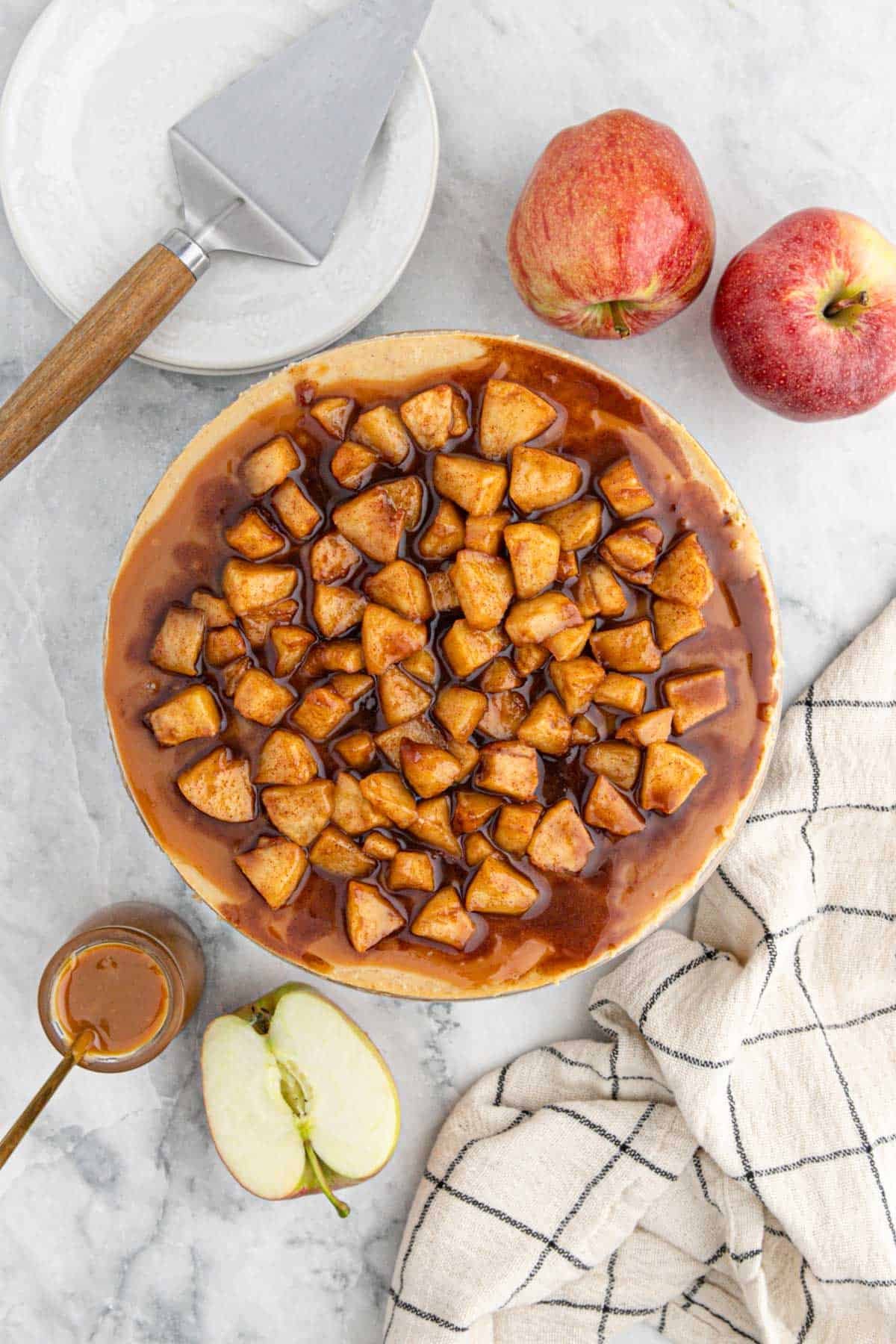 A caramel apple cheesecake topped with cooked cinnamon apples and caramel glaze, ready to serve with fresh apples and plates nearby