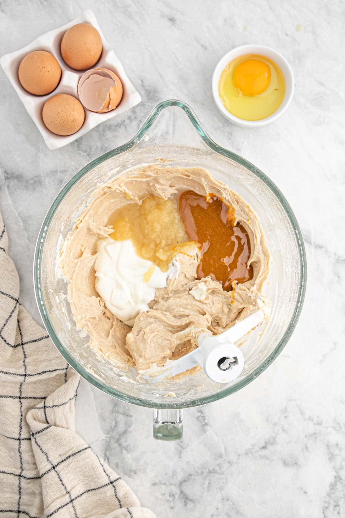 A mixing bowl filled with cheesecake batter, showing added eggs, applesauce, sour cream, and caramel before fully combined