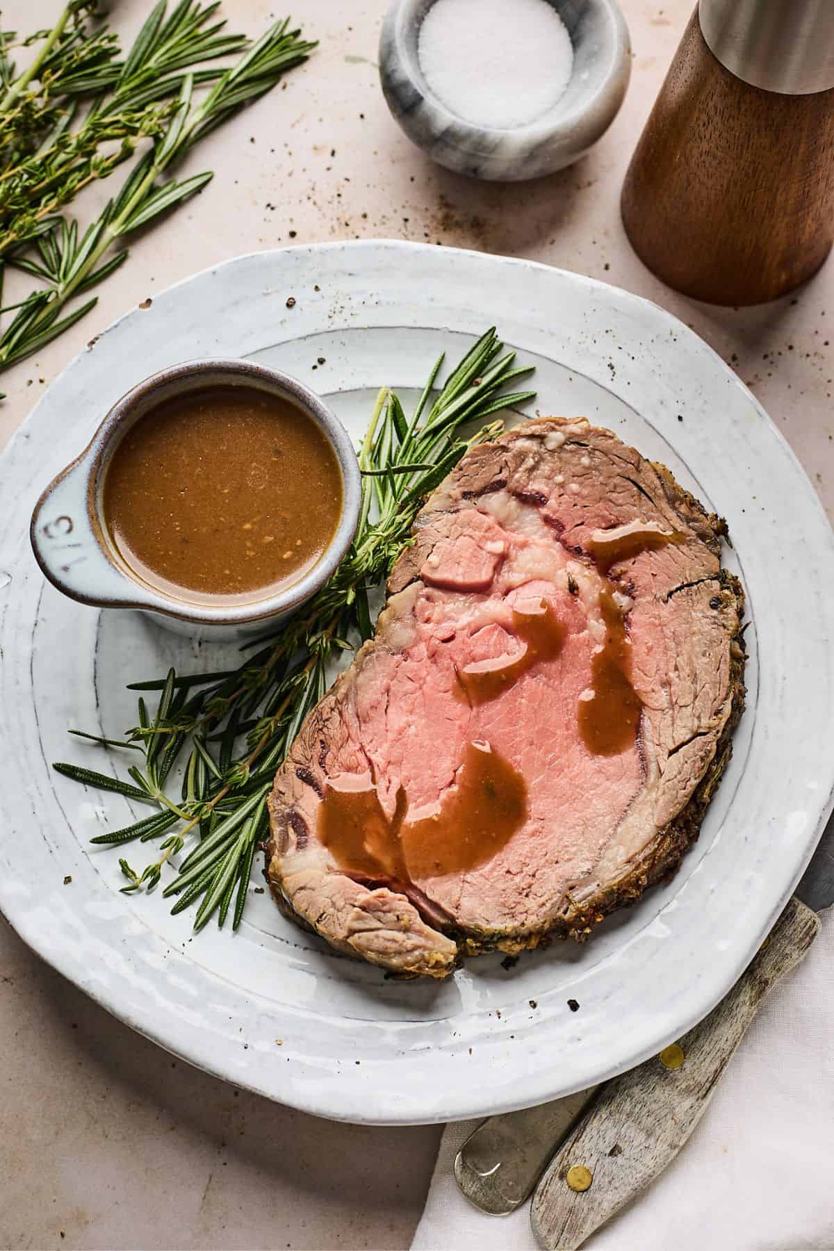 small bowl full of the best au jus recipe next to a slice of prime rib and some rosemary on a white plate
