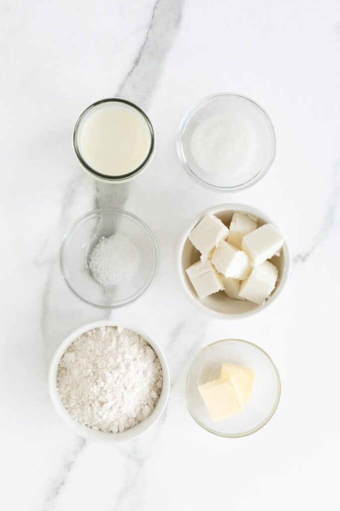 Buttermilk, shortening, butter, salt, leavening and flour in glass bowls on countertop