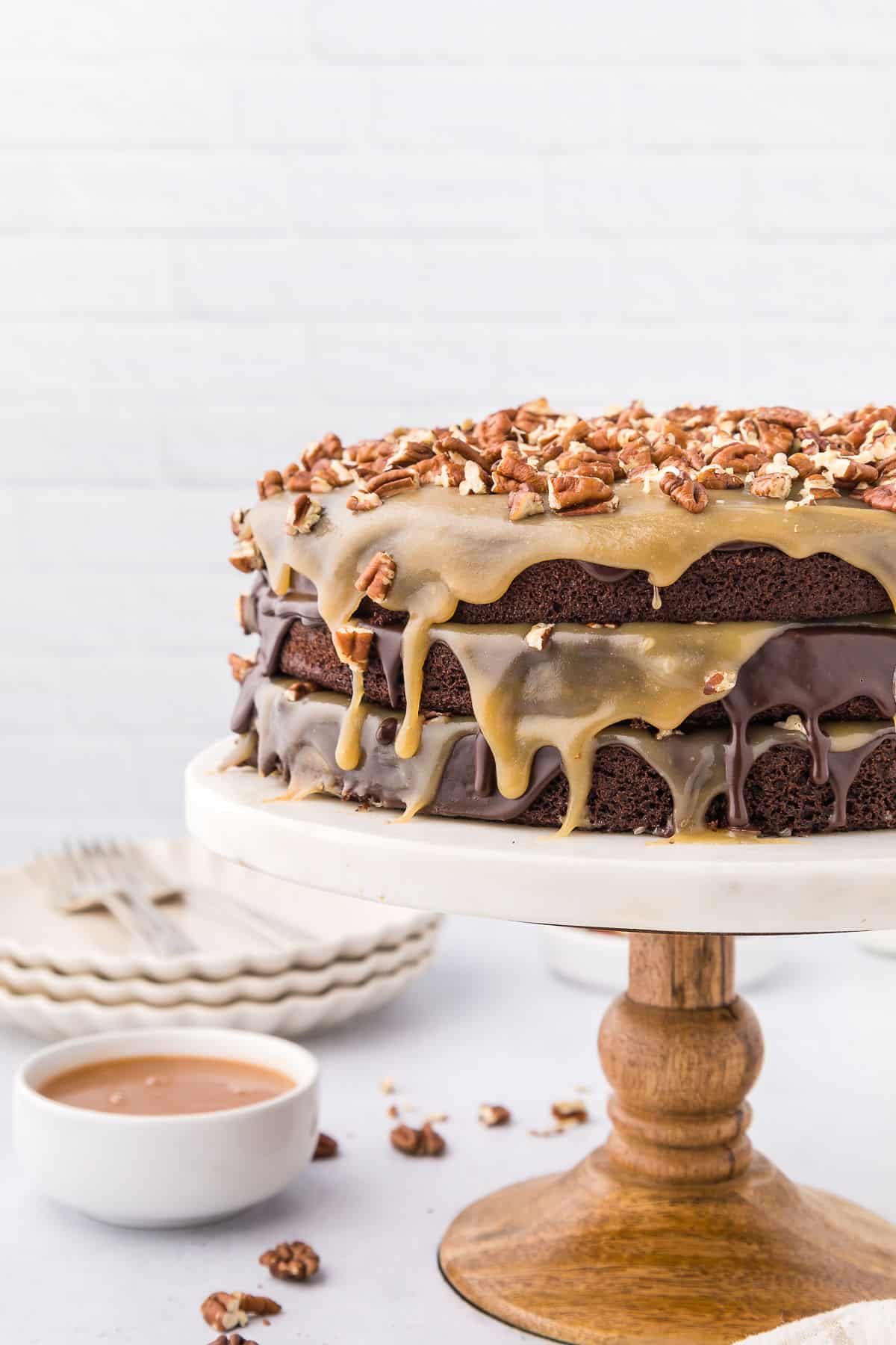 Turtle chocolate cake on a cake stand with pecans and a bowl of caramel sauce next to it