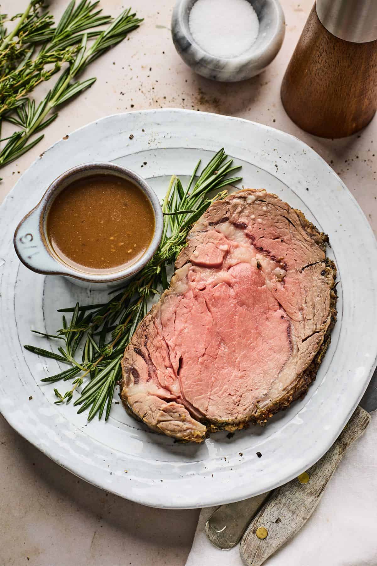 Slice of prime rib recipe with rosemary and a bowl with sauce on a white plate