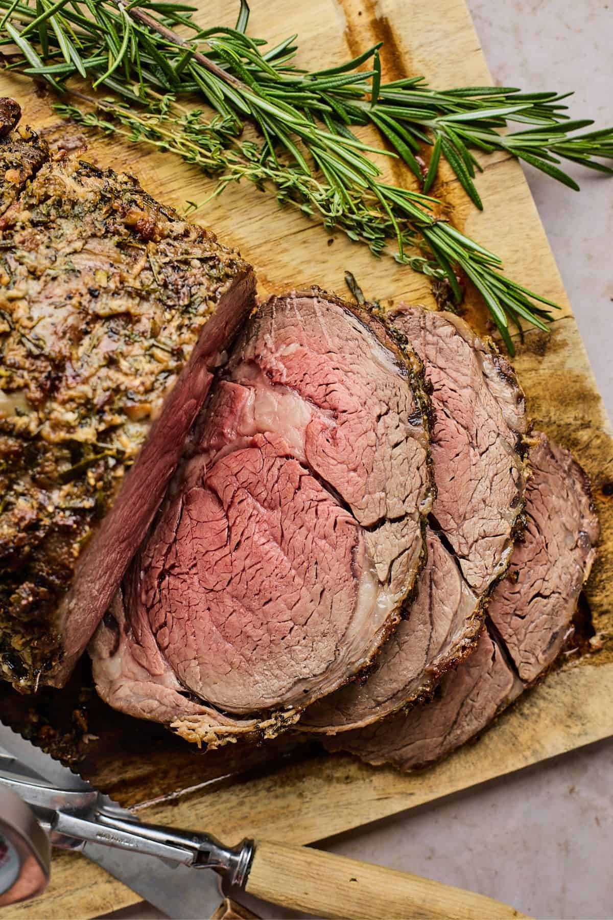 Juicy prime rib recipe sliced on a wooden cutboard with hebrs and a knife next to it