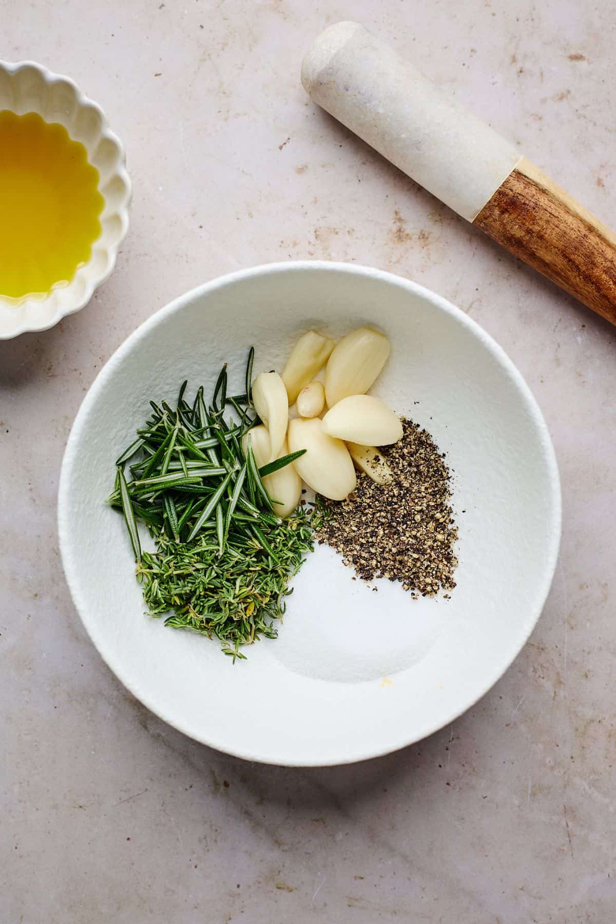 A small bowl with fresh rosemary, thyme, whole garlic cloves, black pepper, and salt, ready to be mixed.