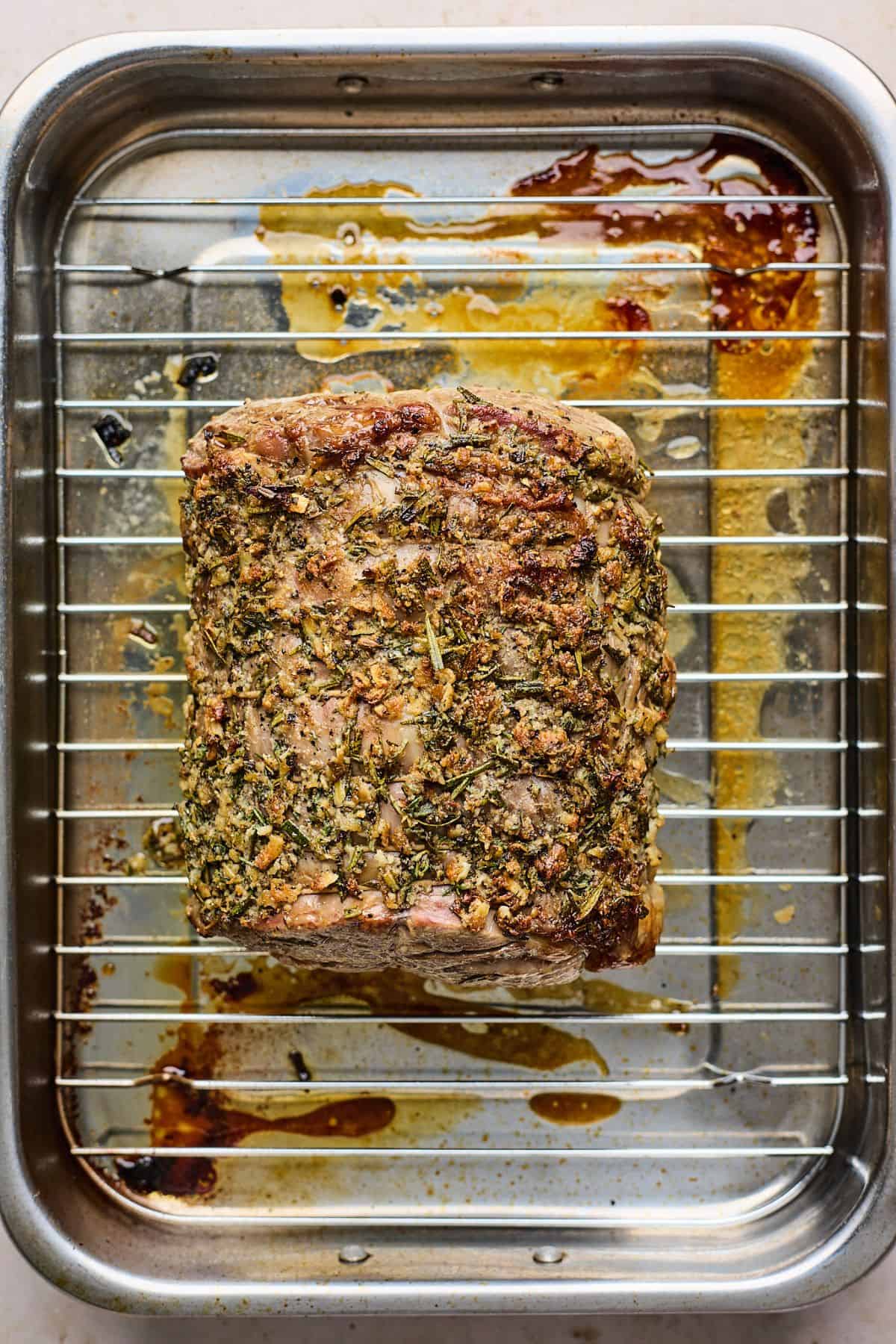 A cooked, herb-crusted prime rib roast resting on a wire rack inside a roasting pan, with golden brown drippings at the bottom.