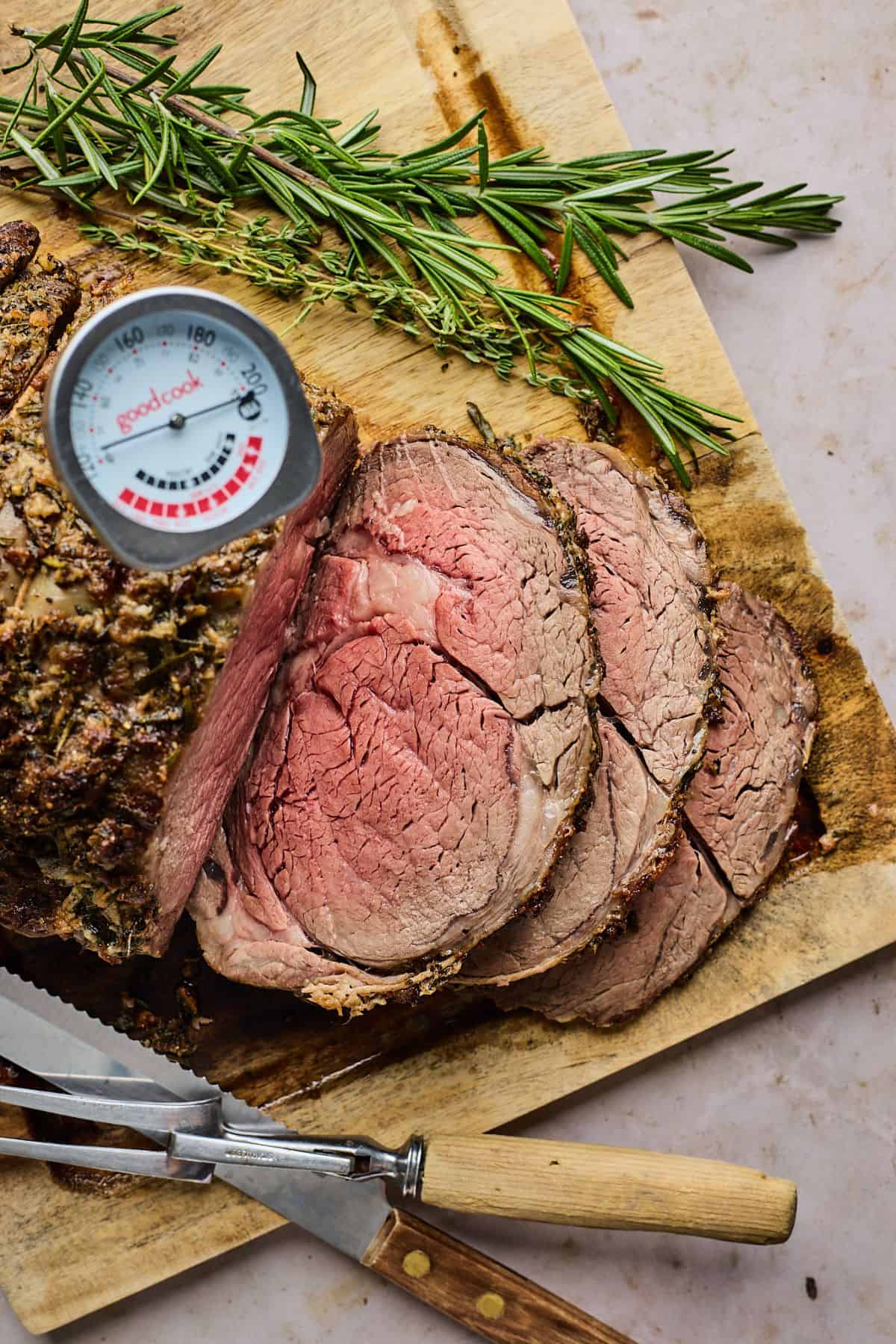 Sliced prime rib roast on a wooden cutting board, accompanied by sprigs of fresh rosemary and thyme, with a meat thermometer inserted into the roast.