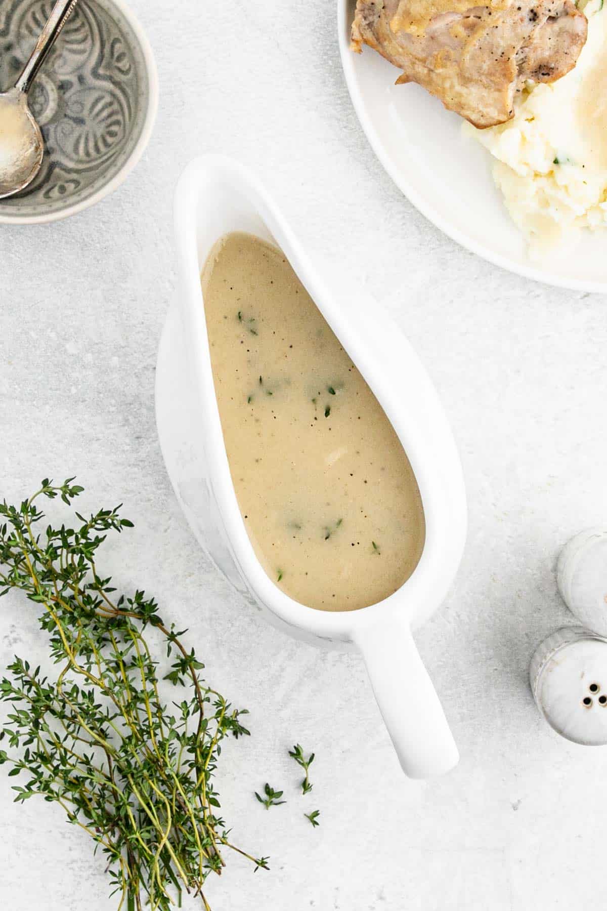 Chicken gravy in a white gravy boat next to fresh thyme and a plate of chicken and mashed potatoes