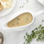 Chicken gravy in a white gravy boat next to fresh thyme and a plate of chicken and mashed potatoes