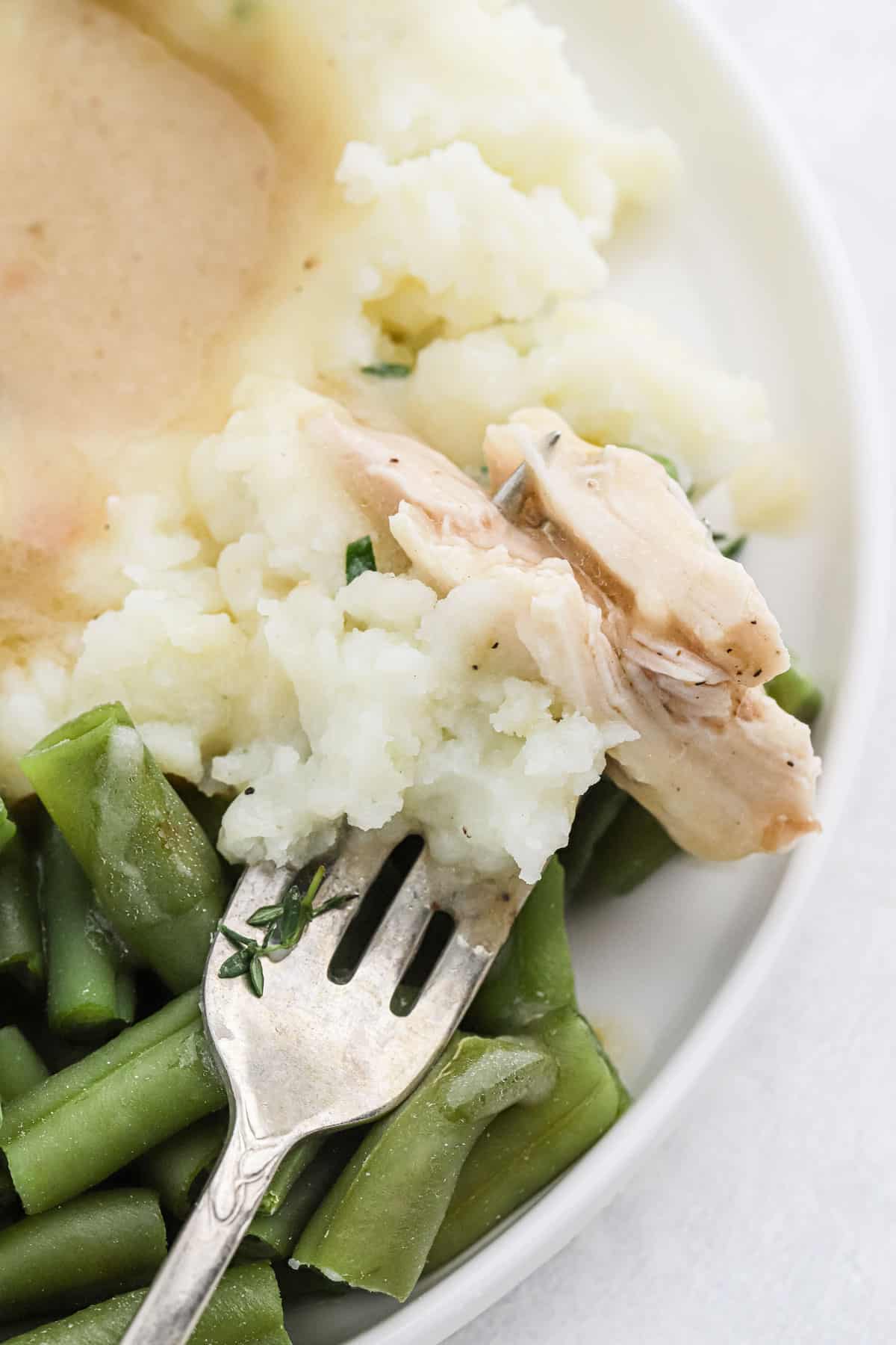 Close up of a fork with some chicken and gravy, mashed potatoes and green beans