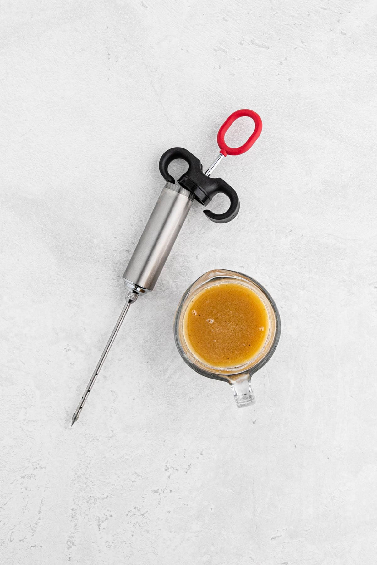 A meat injector filled with marinade, next to a small measuring cup containing marinade