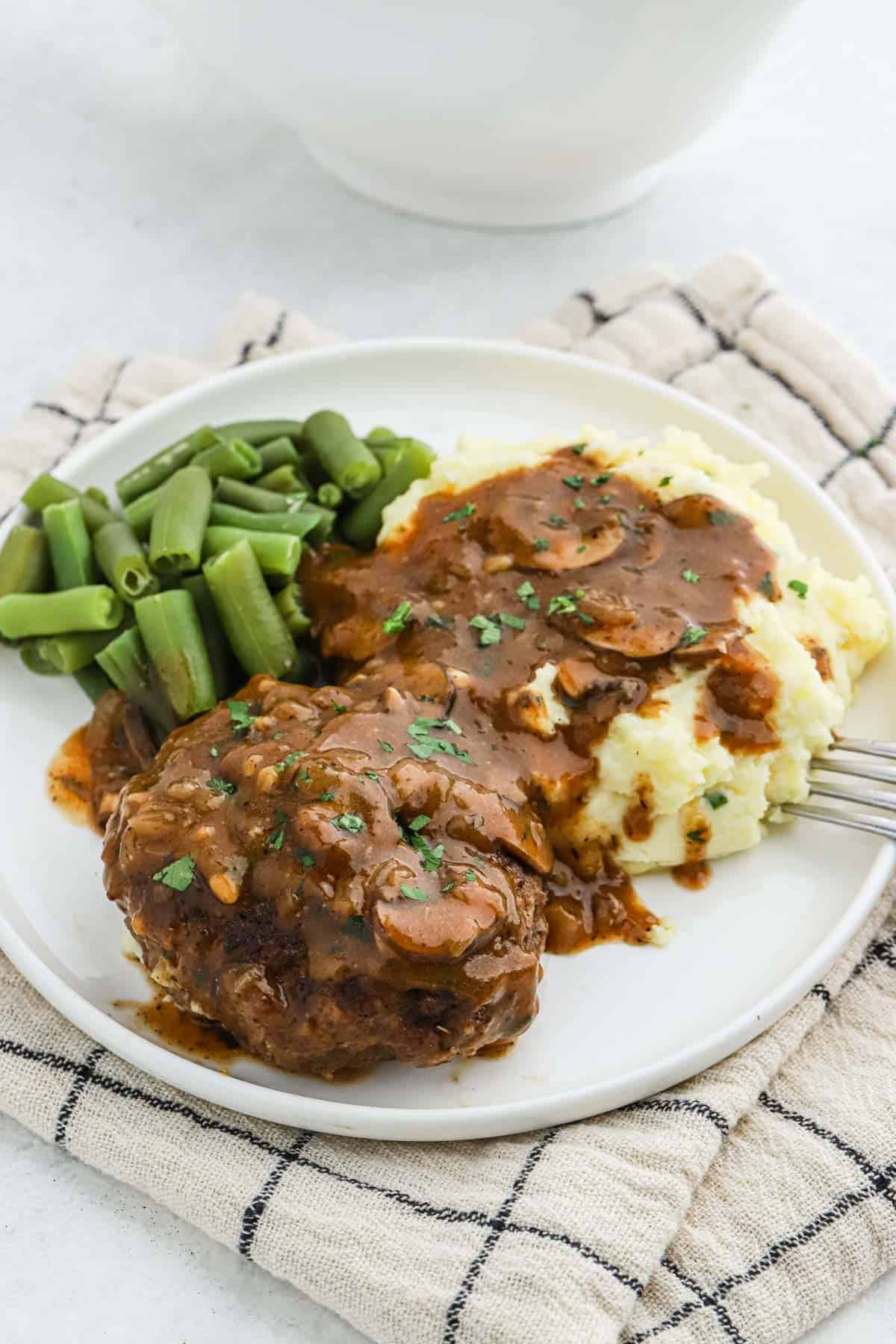 Salisbury steak recipe in a white plate with mashed potatoes and green beans topped with mushroom gravy