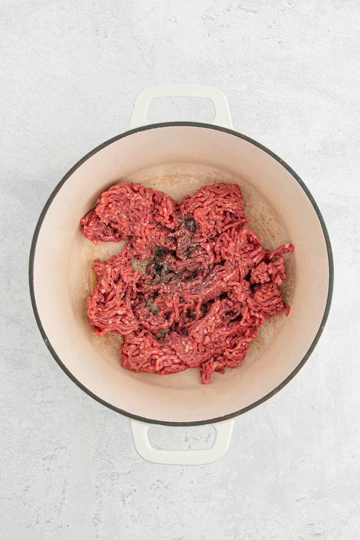 Fresh ground beef seasoned with black pepper and other spices sitting in a cream-colored Dutch oven before cooking