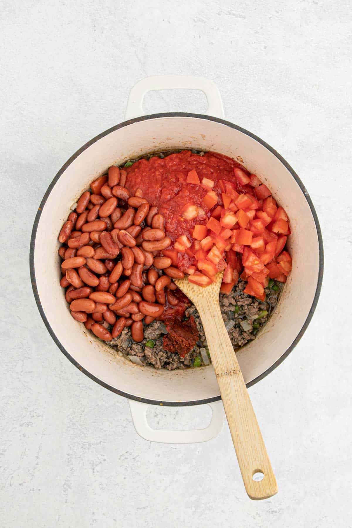 A Dutch oven filled with browned beef, kidney beans, diced tomatoes, and tomato sauce before mixing