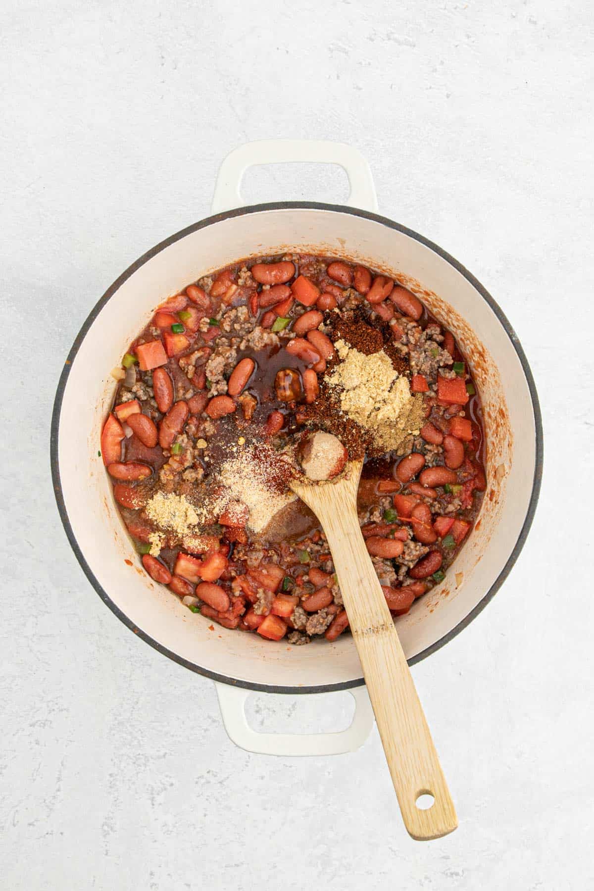 A wooden spoon resting in a Dutch oven filled with chili ingredients, with various dry spices sprinkled on top before stirring