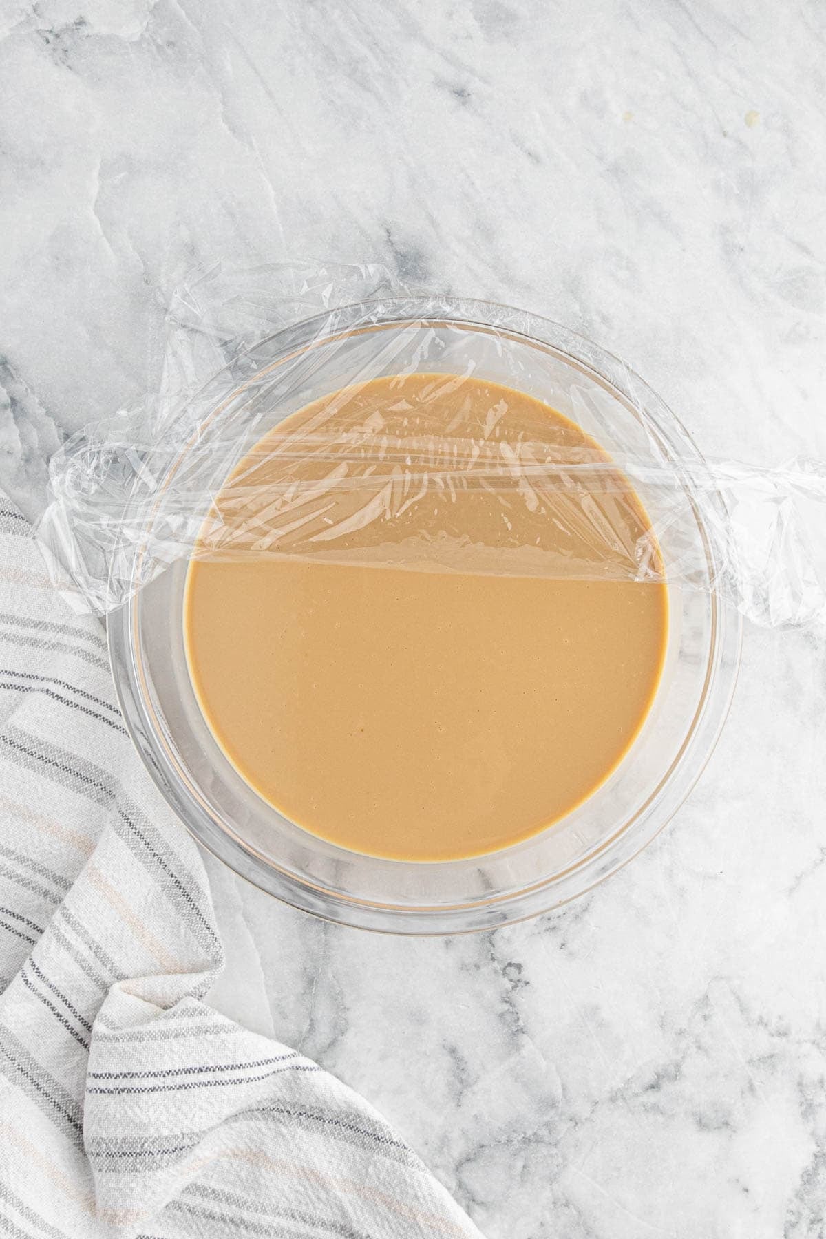 Cookie butter ice cream base covered with plastic wrap in a glass bowl on a marble countertop