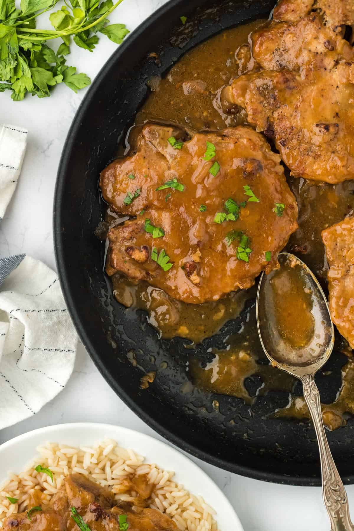 Easy smothered pork chops in a skillet with a spoon against a white background with a pork chop on a white plate with rice