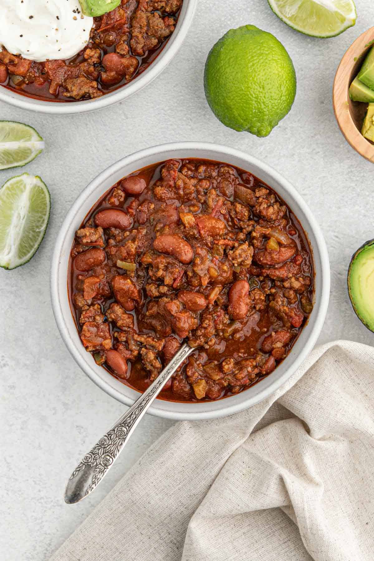 A bowl of best chili on the table without any toppings while limes and avocado are scattered around on the table top.