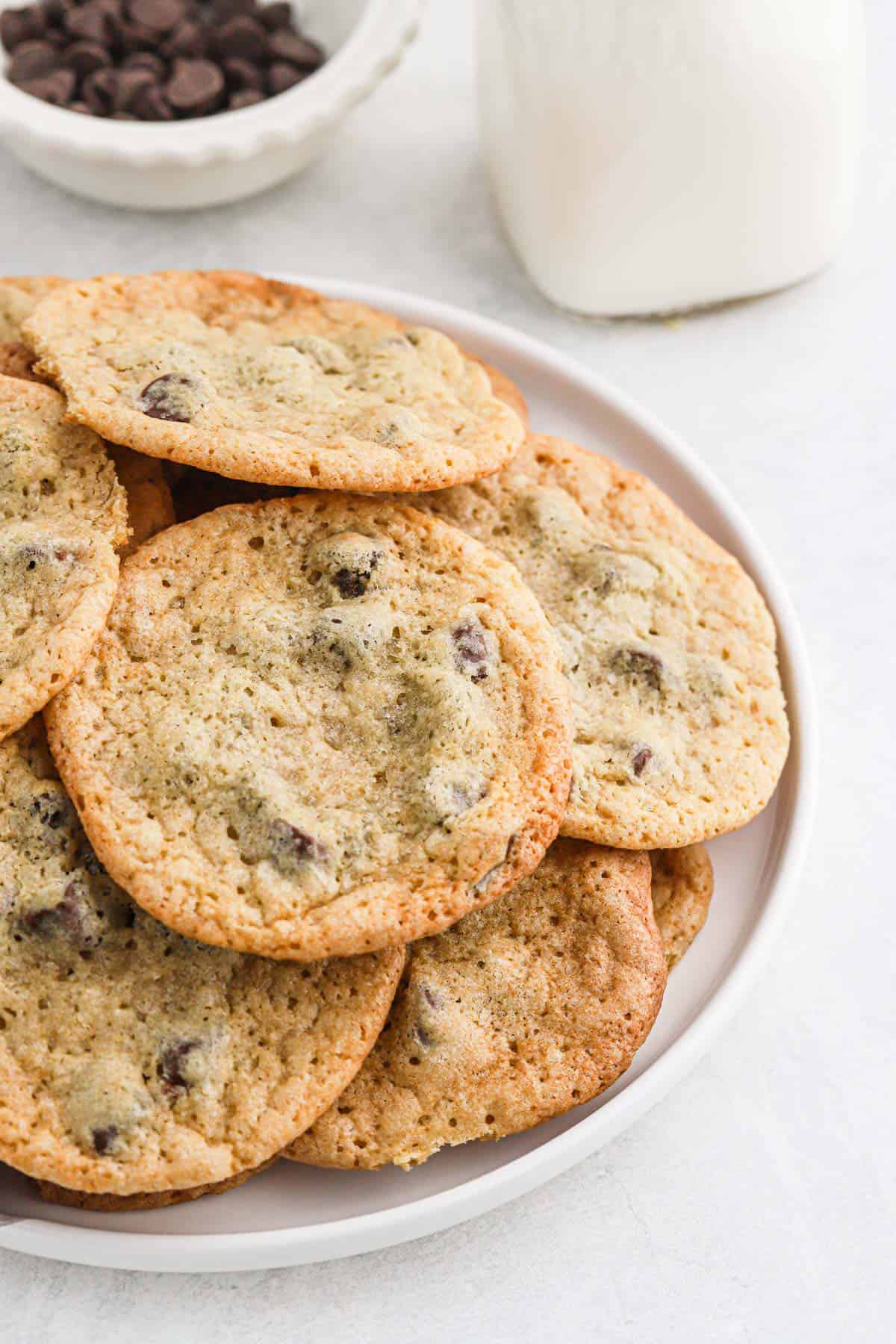 A plate of crispy and thin chocolate chip cookies on the table in front of a jar of milk and a bowl of chocolate chips.