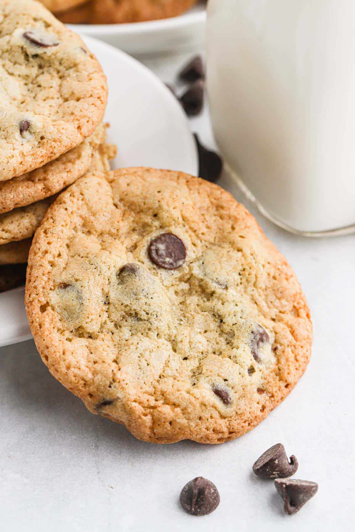 A stack of crispy chocolate chip cookies on a small plate next to a glass of milk with one cookie propped up in front of the stack.