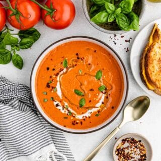 A bowl filled with tomato soup on a white background with tomatoes and parsley in the background
