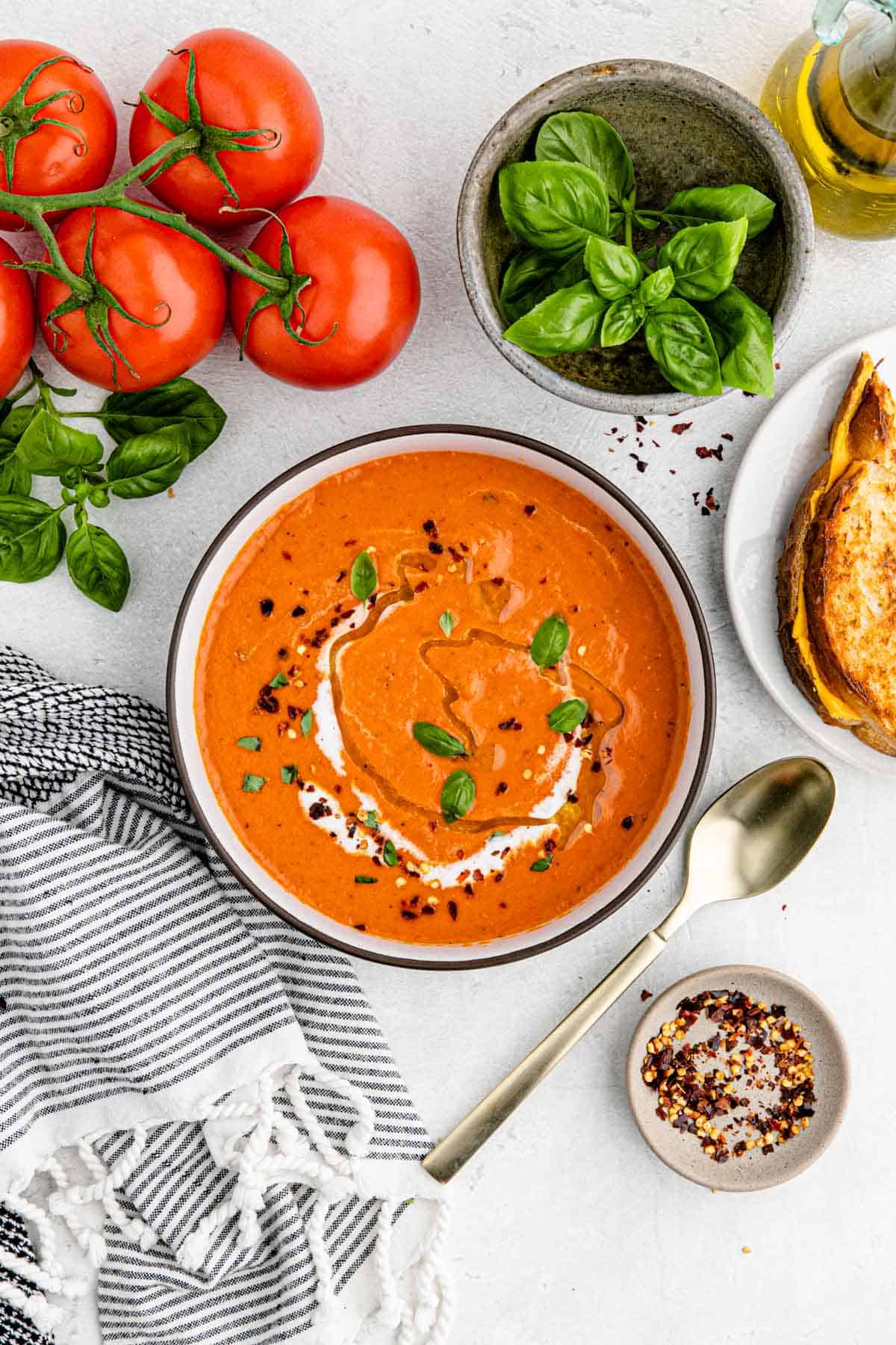 Overhead shot of a bowl of this simple tomato soup with fresh tomatoes, grilled cheese, basil, red pepper flakes and a spoon next to it