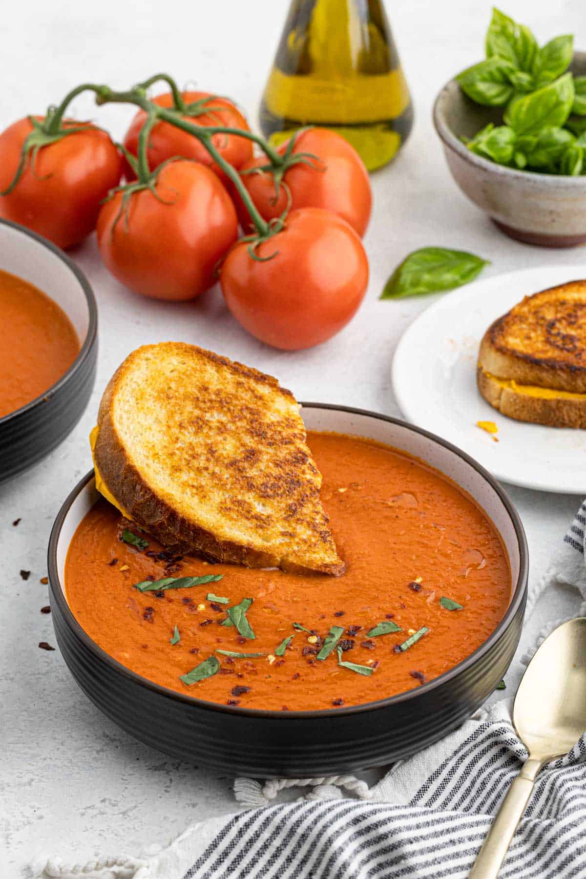 A bowl full of this simple tomato soup with a grilled cheese sandwich inside of it and fresh tomatoes, a spoon and another sandwich next to it
