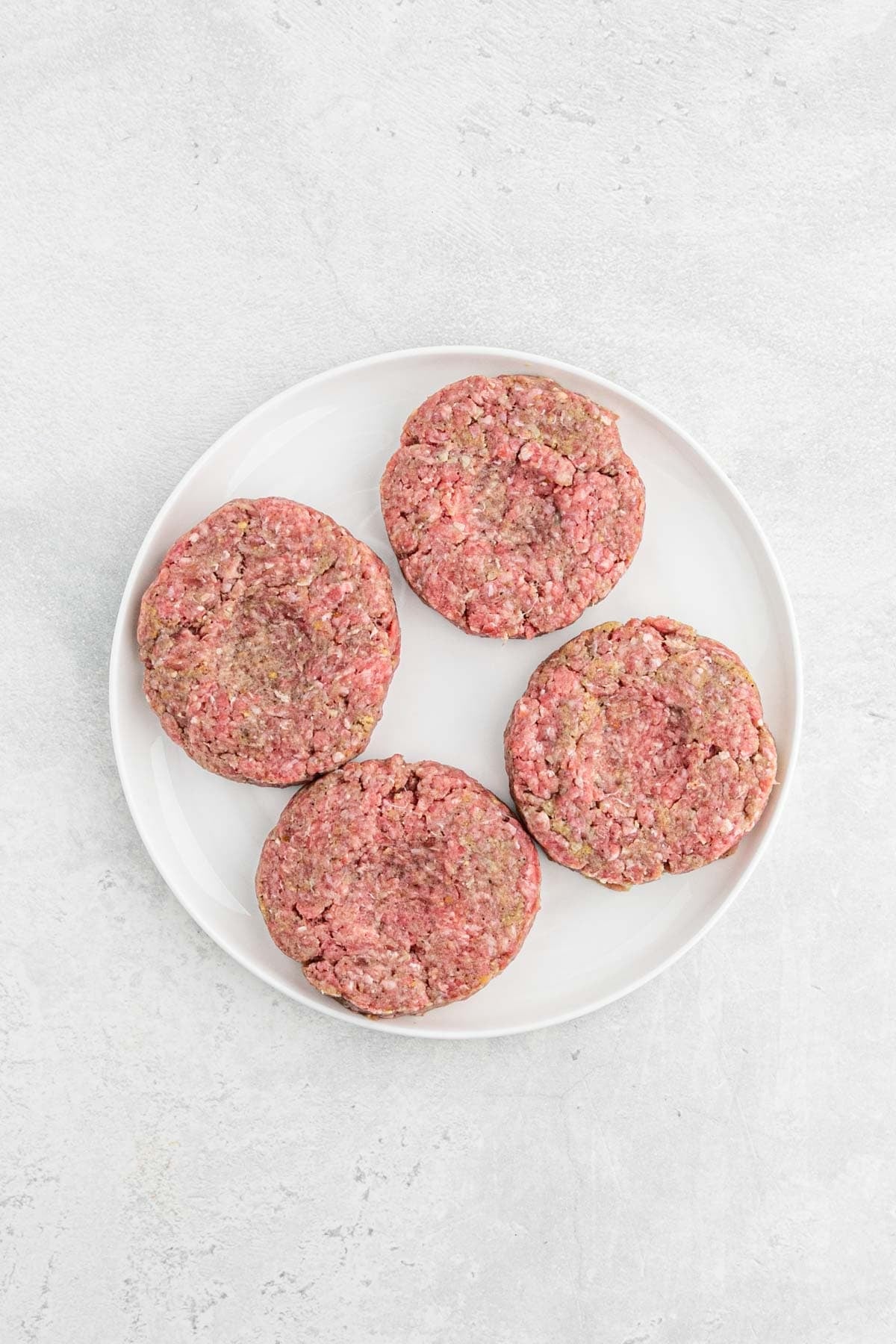 Four raw Salisbury steak patties shaped into thick rounds on a white plate