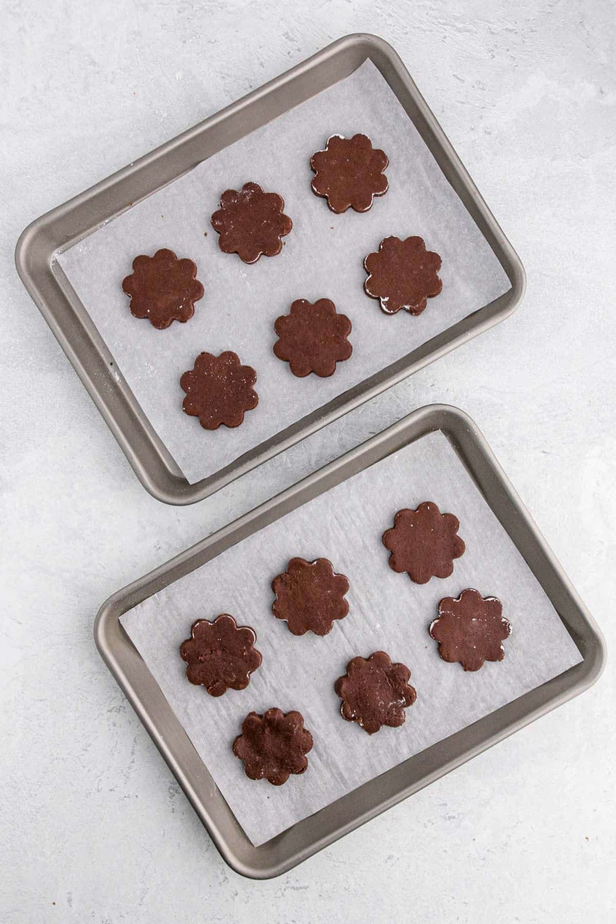 Two baking trays lined with parchment paper, each holding flower-shaped chocolate cookie dough ready to be baked