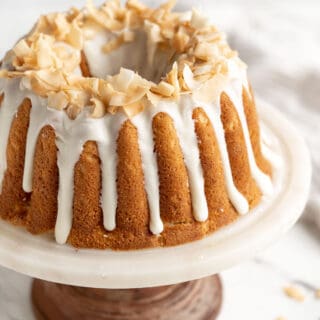 A close up of coconut pound cake on a white and wooden cake stand on a white background
