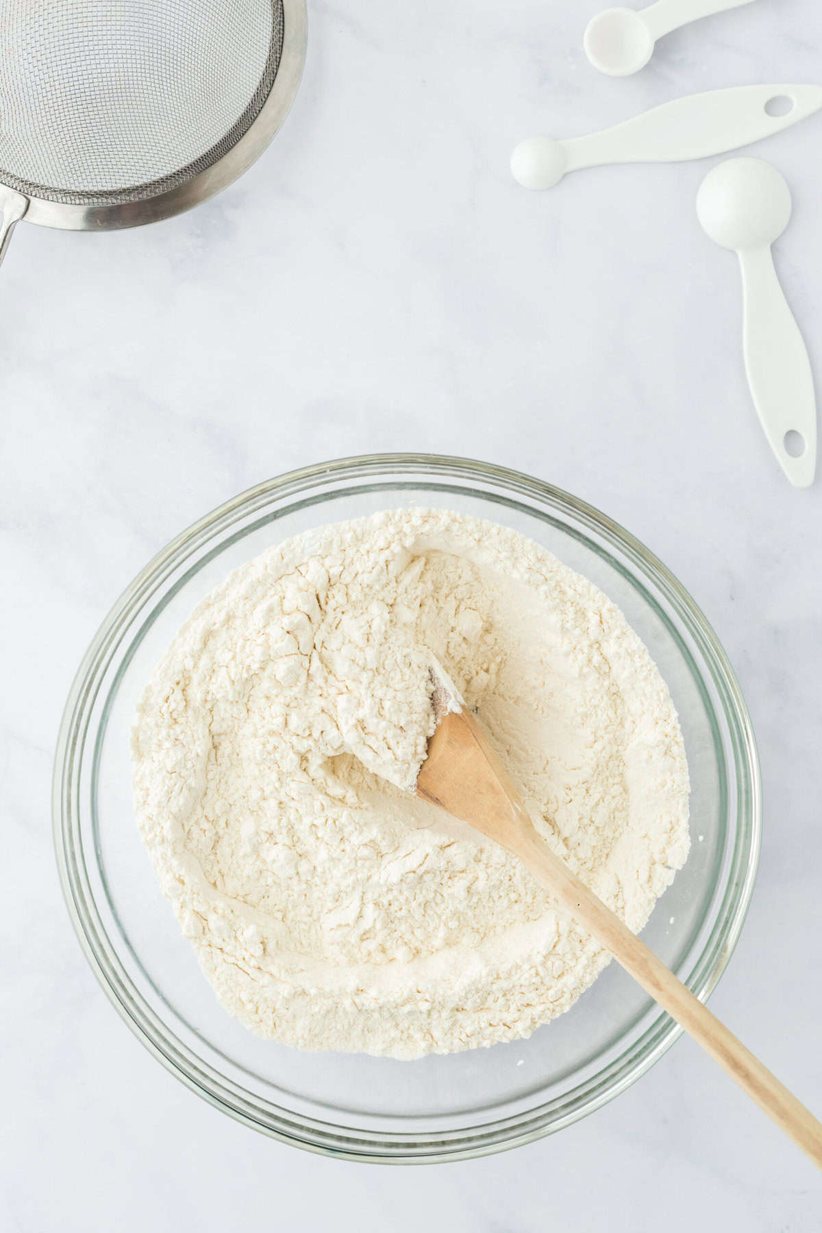 Dry ingredients in a glass bowl with a wooden spoon mixing together on white background