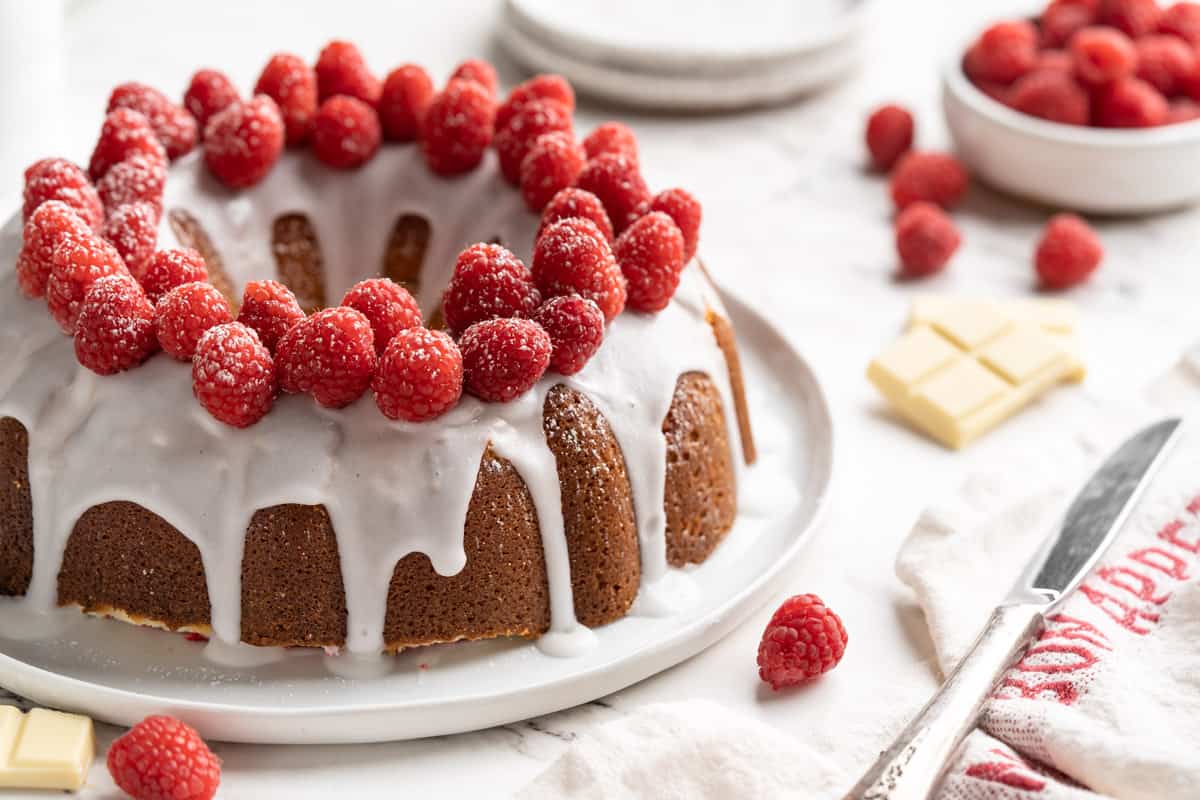 White chocolate raspberry bundt cake on a white plate with raspberries, white chocolate and a knife next to it