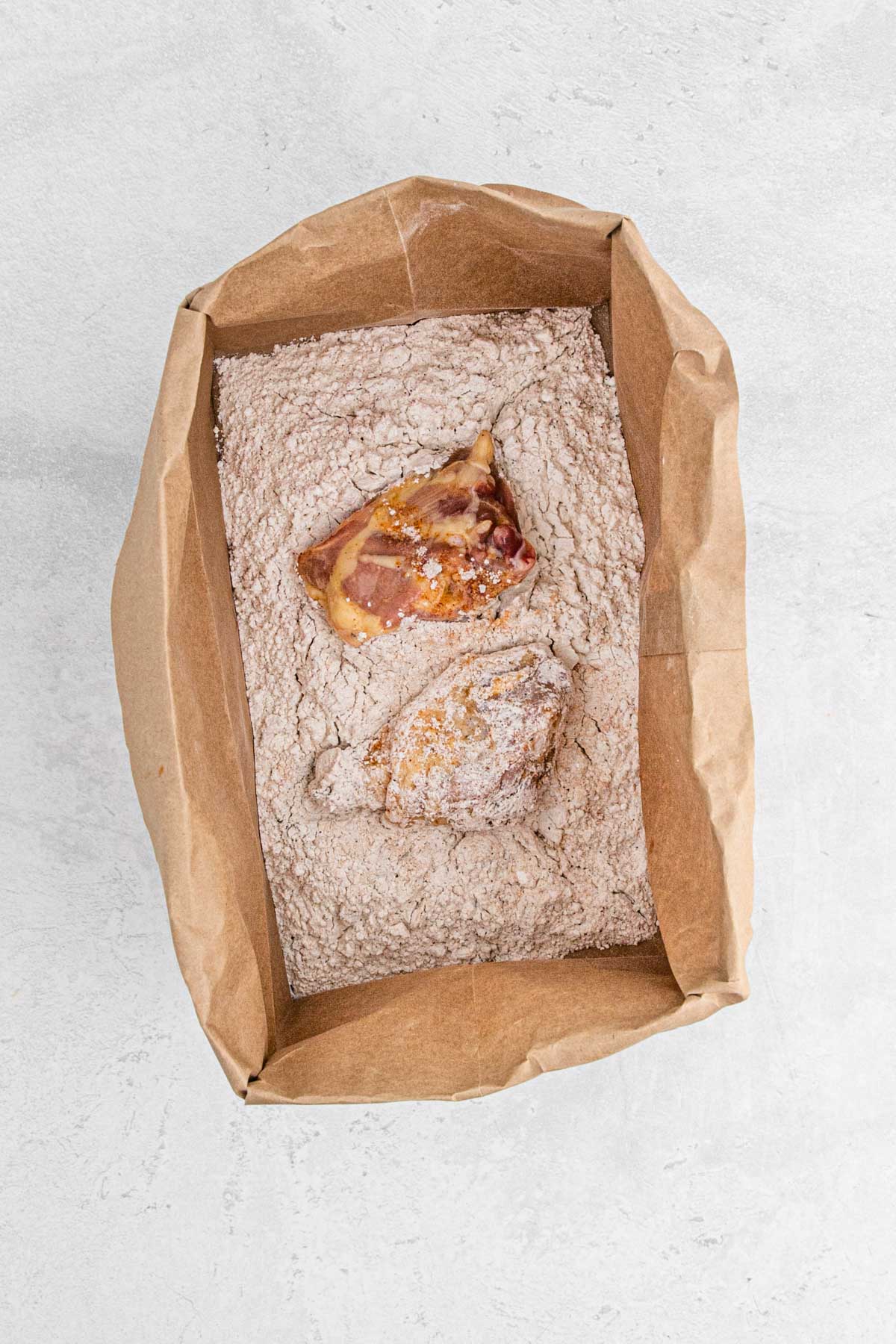 Raw, marinated chicken being coated in the flour and spice mixture inside the paper bag