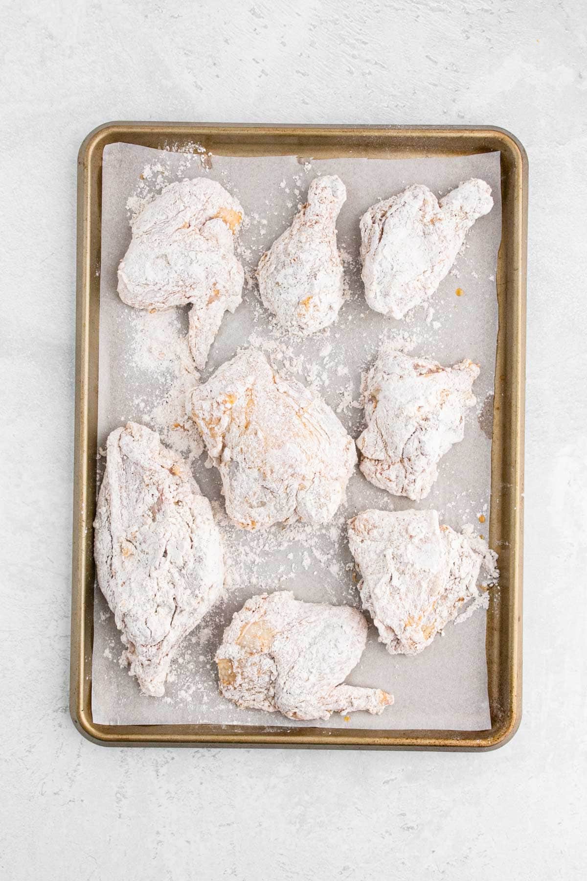 Several pieces of raw chicken, fully dredged and resting on a parchment-lined baking tray