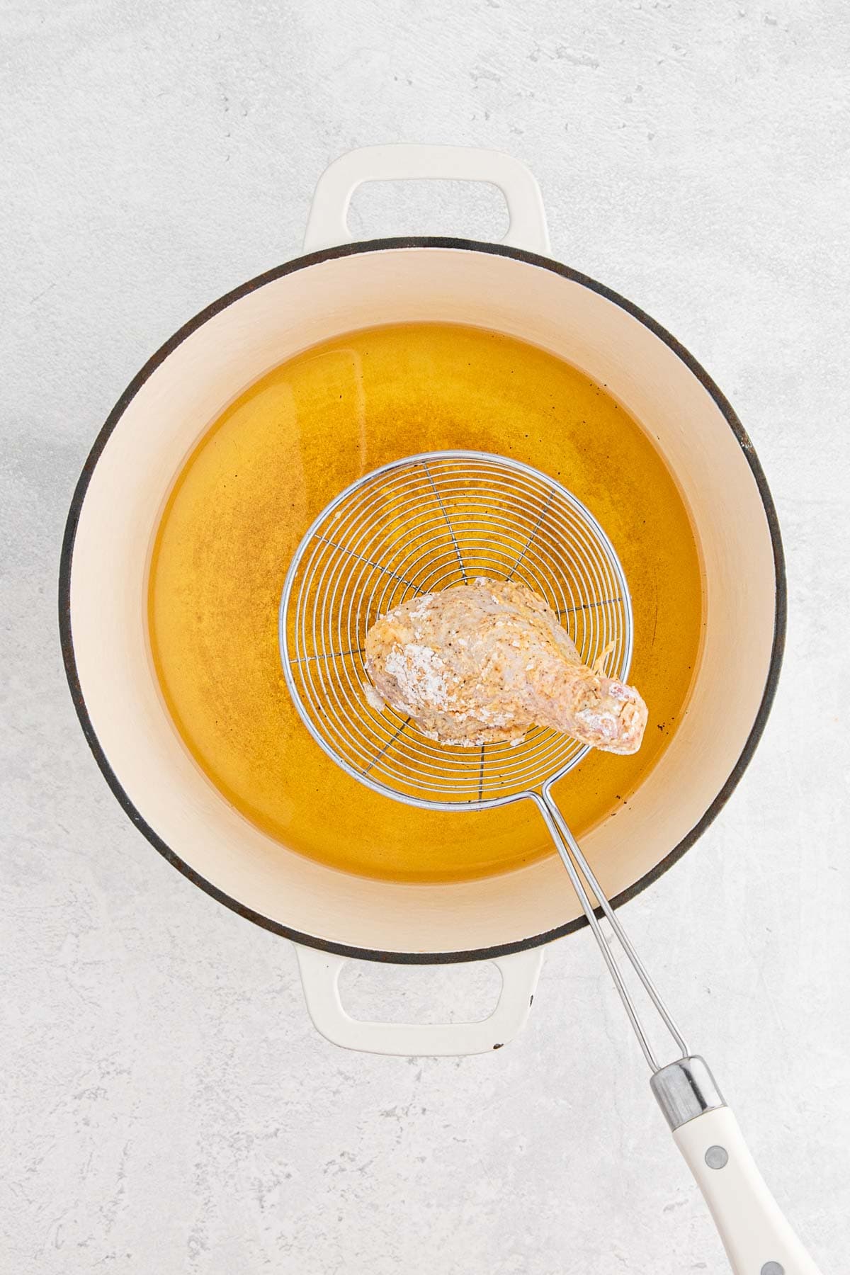 A piece of coated raw chicken held above a pot of hot oil with a spider strainer