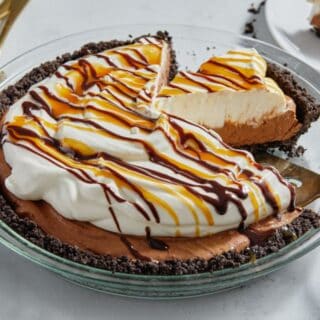 Chocolate mousse pie drizzled with caramel and chocolate sauce in a glass dish, with one slice partially lifted out by a cake server