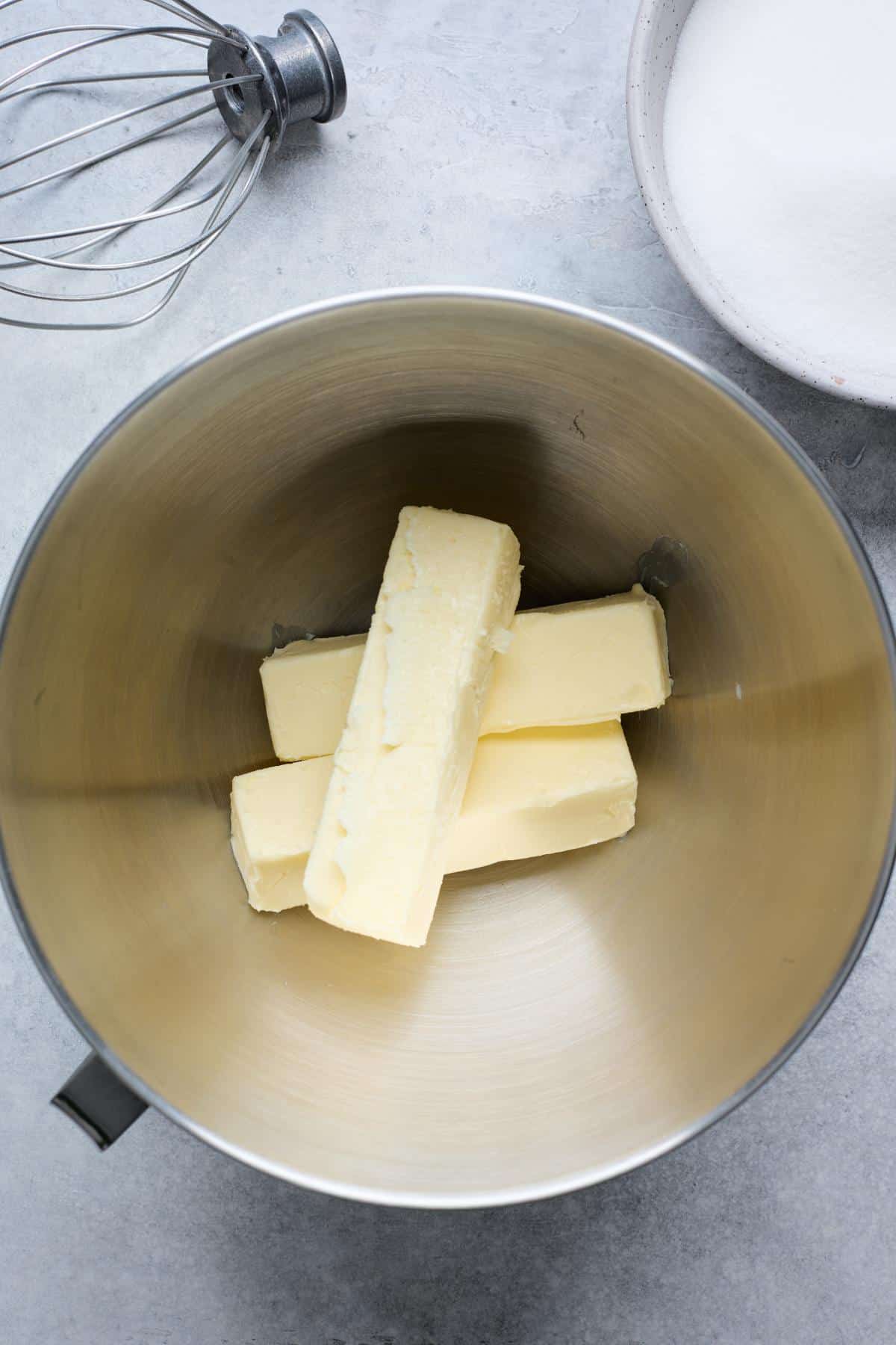 Butter sticks in a metal mixing bowl, with a whisk attachment and sugar bowl visible in the background