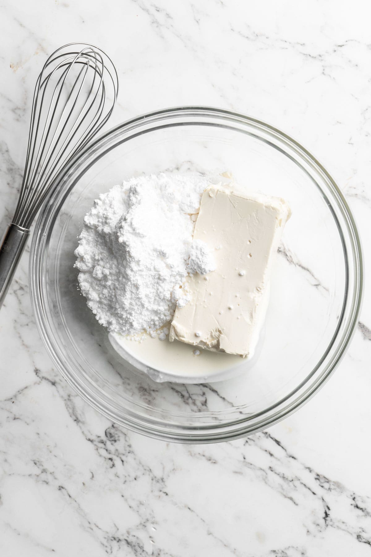 Cream cheese and powdered sugar with milk being added to a glass bowl on white countertop