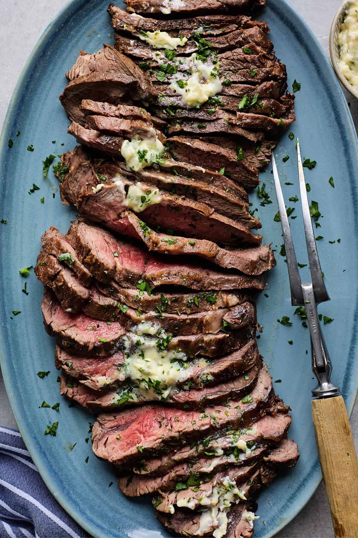 London broil smothered with herb butter sliced on a blue plate with a carving fork next to it
