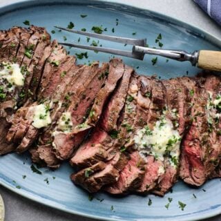 London broil smothered with herb butter sliced on a blue plate with a carving fork next to it