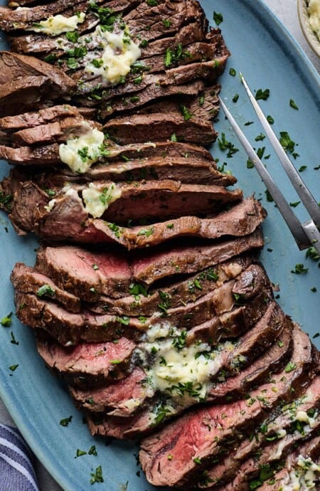 London broil smothered with herb butter sliced on a blue plate with a carving fork and more herb butter in a bowl next to it