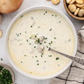 Overhead shot of oyster stew in a white bowl with a silver spoon, with onions, oyster crackers and chives in bowls next to it