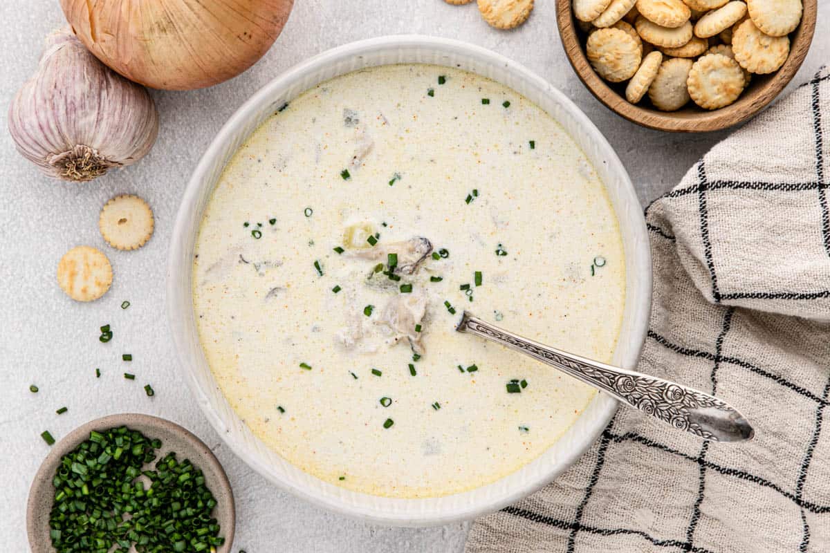 Overhead shot of oyster stew in a white bowl with a silver spoon, with onions, oyster crackers and chives in bowls next to it
