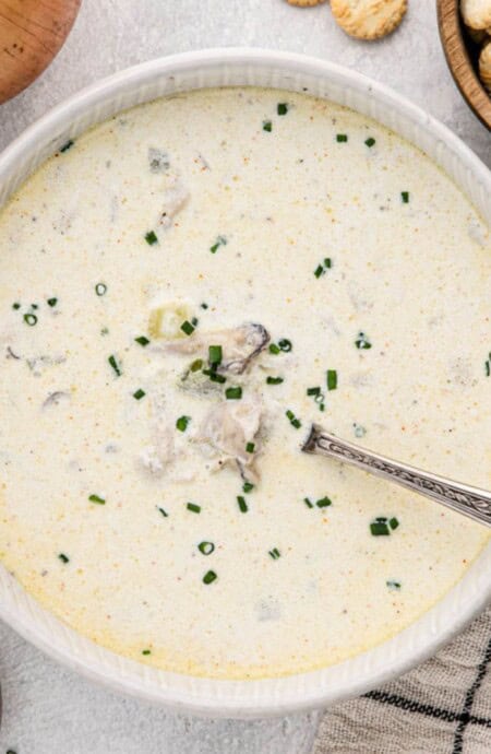 Overhead shot of oyster stew in a white bowl with a silver spoon, with oyster crackers and chives in bowls next to it