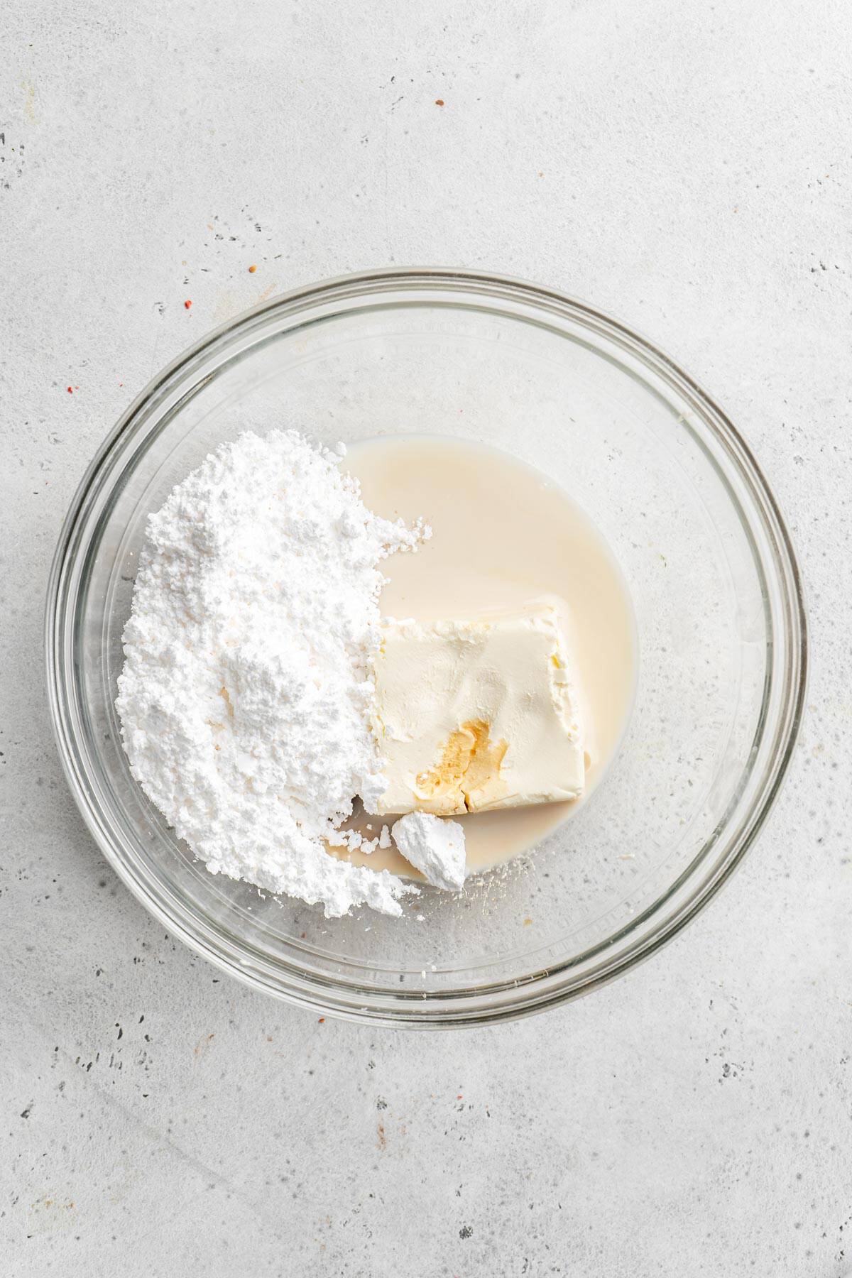 Cream cheese, milk and powdered sugar in a glass bowl on white countertop