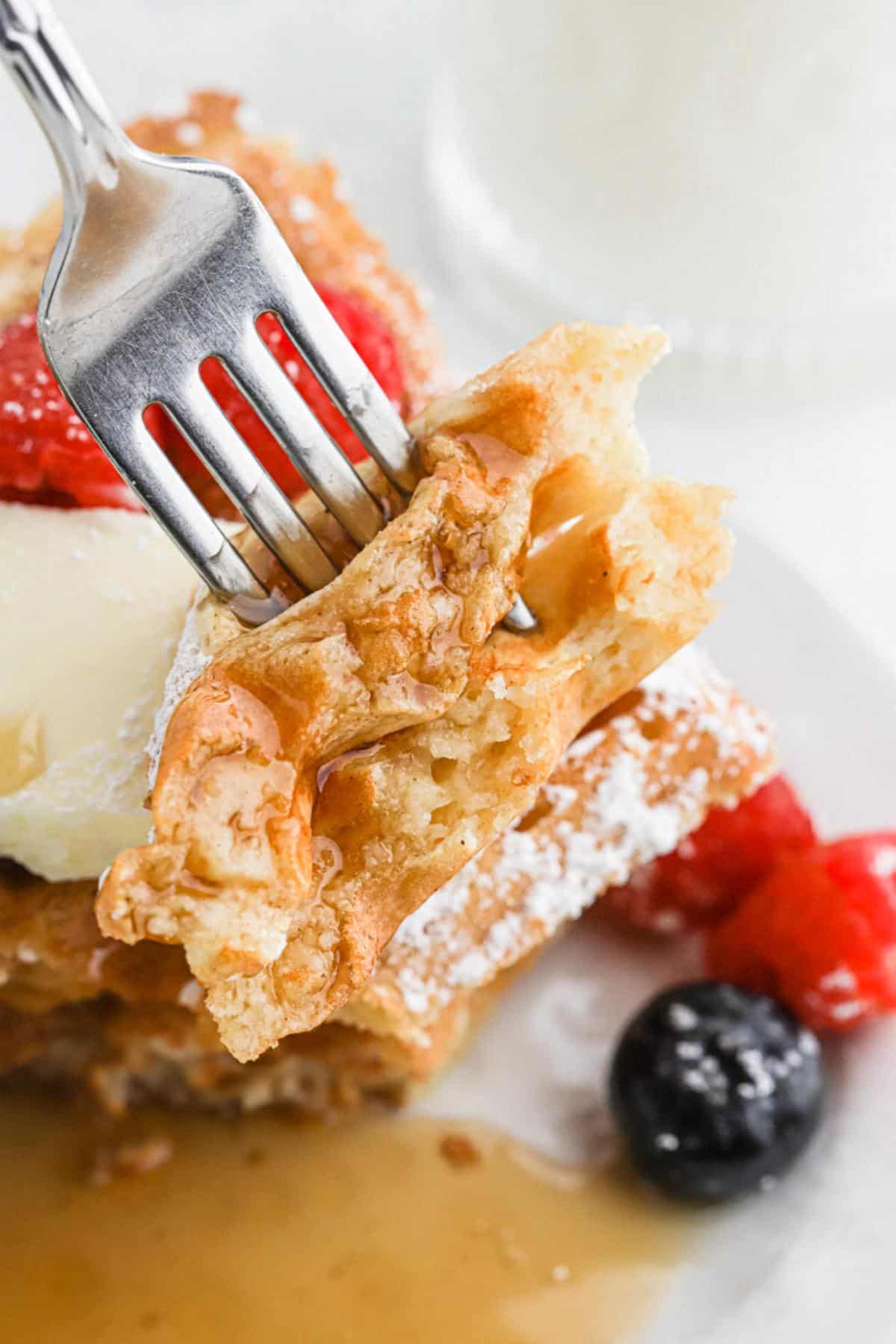 Fork lifting a piece of buttermilk waffles topped with syrup and powdered sugar, with berries in the background