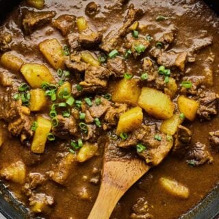 Curry goat in a large dutch oven with potatoes and garnished with parsley, with a wooden spoon stirring it